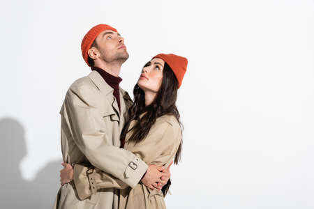 trendy couple in beanie hats and trench coats hugging and looking up on whiteの写真素材