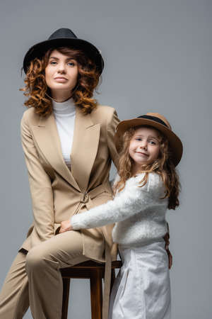elegant mother and daughter in white and beige outfits posing on chair isolated on grayの写真素材