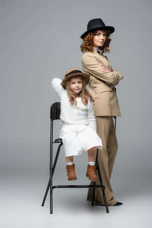elegant mother and daughter in white and beige outfits and hats posing on chair on gray backgroundの写真素材