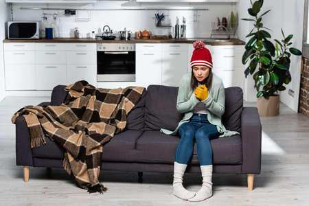 shivering woman in warm hat, gloves and socks sitting on sofa near plaid blanket in kitchenの写真素材