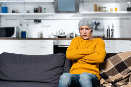 shivering man in knitted sweater and hat sitting on sofa with crossed arms in cold kitchenの写真素材