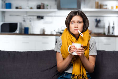 ill woman with thermometer in mouth holding cup of warm tea in kitchenの写真素材
