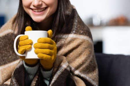 cropped view of woman wearing knitted gloves, wrapped in plaid blanket, holding cup of warm tea at...