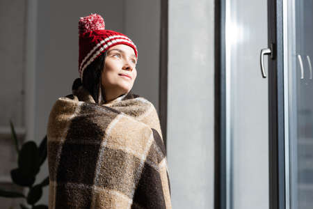 dreamy woman in knitted hat, wrapped in warm plaid blanket, looking away while standing near window...