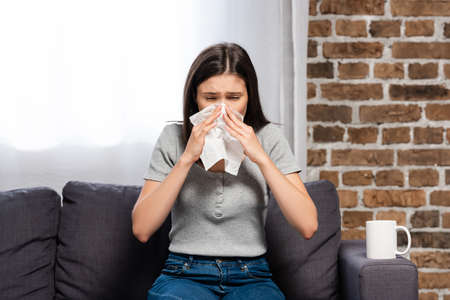 diseased woman sneezing in paper napkin while sitting on sofa near cup of warming beverageの写真素材