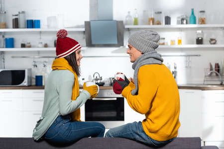 side view of young couple in warm hats, scarfs and gloves holding cups with tea in kitchenの写真素材