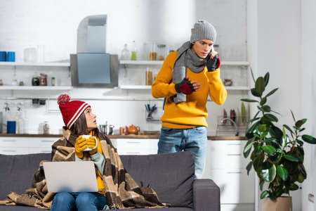 cold woman in warm hat and gloves sitting on sofa under plaid blanket while worried boyfriend...