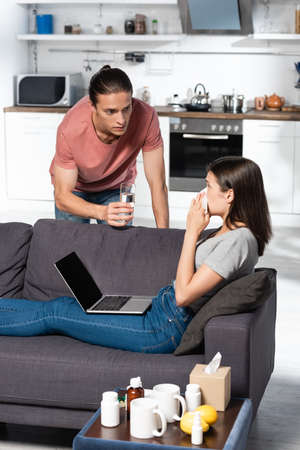 young man giving glass of water to sick woman sitting on sofa with laptop and sneezing in paper napkinの写真素材