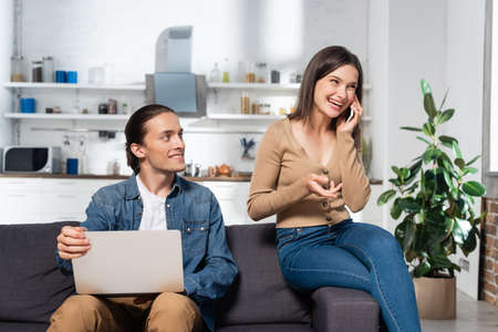 excited woman talking on smartphone near man using laptop on sofa in kitchen