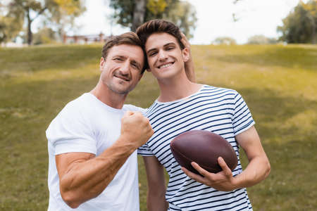 proud father showing clenched fist and standing near teenager son with rugby ballの写真素材