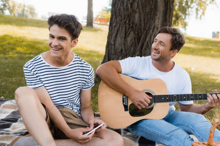 selective focus of teenager boy holding smartphone near father playing acoustic guitarの写真素材