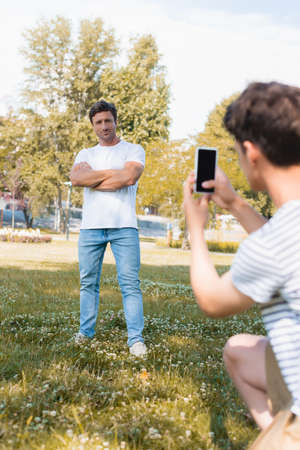 selective focus of man standing with crossed arms while teenager boy taking photo and holding smartphone in parkの写真素材
