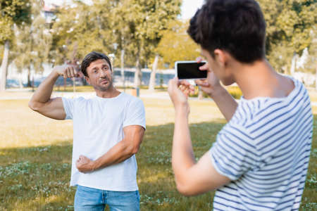 selective focus of strong father posing while teenager boy taking photo and holding smartphone in parkの写真素材