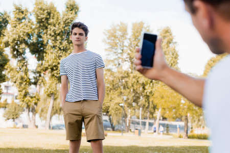 selective focus of teenager boy standing with hands in pockets while father taking photo on smartphone in parkの写真素材