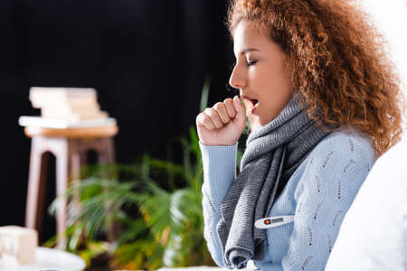 Selective focus of curly woman in scarf using thermometer and coughing at homeの写真素材