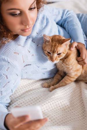 Selective focus of woman petting cat while using smartphone on bedの写真素材
