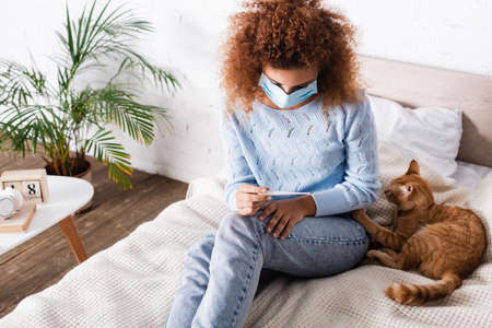 Diseased woman in medical mask holding thermometer while sitting near cat on bedの写真素材