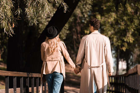back view of couple in trench coats holding hands and walking on wooden bridge in autumnal parkの写真素材