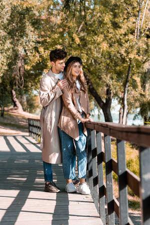 man touching shoulders of woman in hat and trench coat standing on wooden bridge in autumnal parkの写真素材