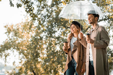 couple in trench coats standing under umbrella and holding paper cups with coffee to goの写真素材