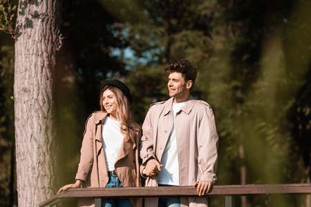 stylish couple in trench coats looking away and holding hands in parkの写真素材