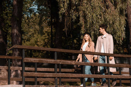 couple in trench coats holding hands and walking on wooden bridgeの写真素材