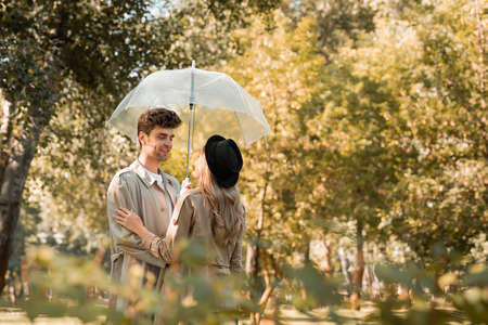 selective focus of woman in hat and man in trench coat standing under umbrella in autumnal parkの写真素材