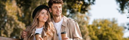 panoramic shot of blonde woman in hat and man in trench coat holding paper cups in autumnal parkの写真素材