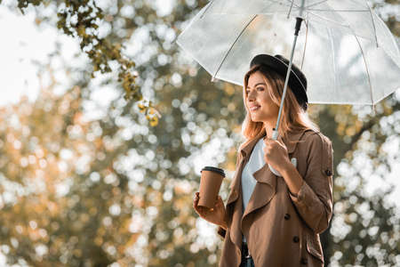 woman in trench coat and hat standing under umbrella and holding paper cup in autumnal park