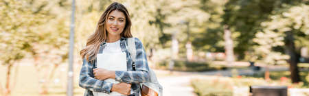 panoramic shot of woman in plaid shirt holding laptop and looking at camera in park