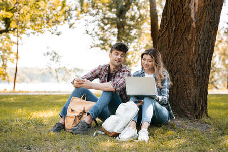 man holding smartphone and looking at laptop near woman in parkの写真素材