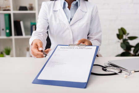 cropped view of doctor holding insurance claim form near stethoscope on desk