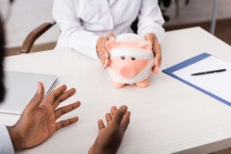 cropped view of african american patient gesturing with hands near doctor touching piggy bank,...