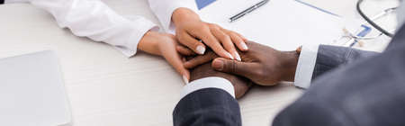 partial view of doctor touching hands of african american man near clipboard and pen on desk,...