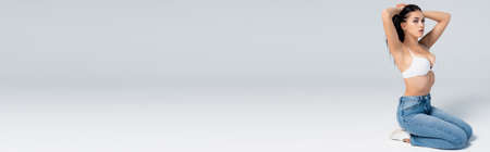 panoramic concept of brunette woman in bra and jeans sitting and touching hair on grayの写真素材