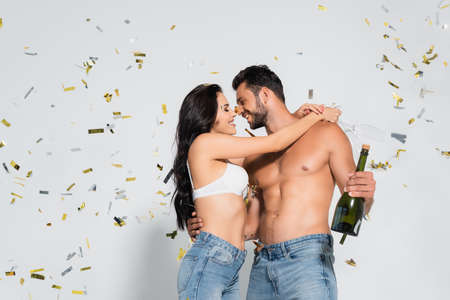 brunette woman in bra hugging muscular man with bottle of champagne on gray with confettiの写真素材