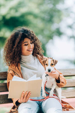 curly woman in autumn outfit sitting on bench in park with book and jack russell terrier dogの写真素材