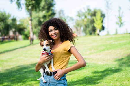 young woman in summer outfit standing with hand in pocket while holding jack russell terrier dog in parkの写真素材