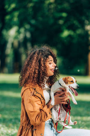 Curly woman in raincoat holding jack russell terrier with leash in parkの写真素材