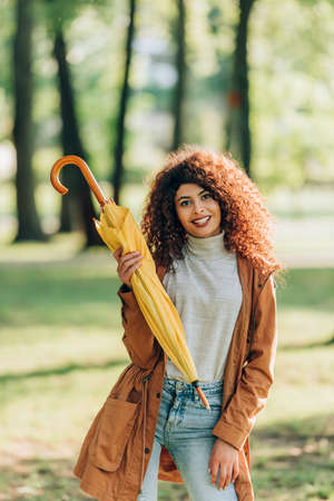 Young woman in raincoat holding umbrella and looking at camera in parkの写真素材