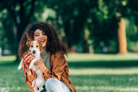 Young woman in raincoat holding jack russell terrier and looking at camera in parkの写真素材