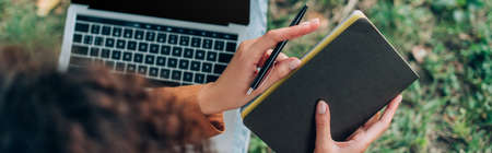 Website header of teleworker holding notebook and pen while using laptop on grass in park