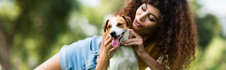 horizontal orientation of curly woman cuddling jack russell terrier dog while resting in parkの写真素材