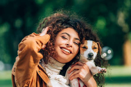 Selective focus of young curly woman with hand near head holding jack russell terrier outdoorsの写真素材