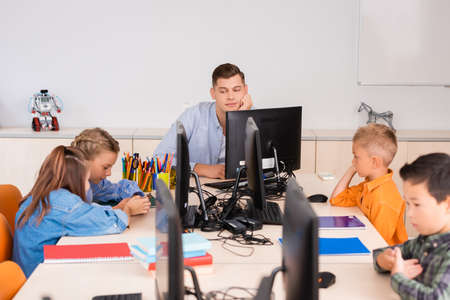 Selective focus of teacher looking at multiethnic pupils near computers in stem school