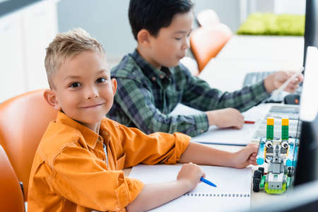 Selective focus of schoolboy looking at camera near robot and asian friend in stem school