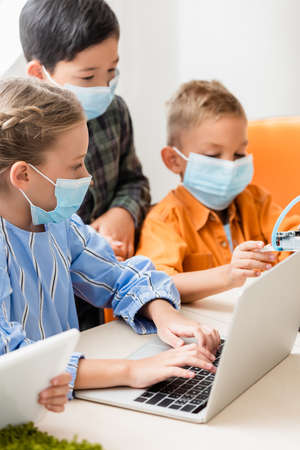 Selective focus of multicultural children in medical masks using gadgets while programming robot in...