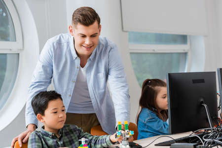 Selective focus of teacher standing near asian schoolboy holding robot and computer in classroom