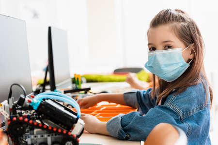 Selective focus of schoolgirl in medical mask sitting near robot and computers in classroom