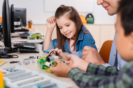 Selective focus of teacher sitting near multiethnic pupils and parts of robot in classroom